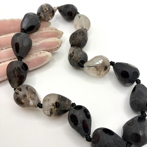 May include: A necklace featuring a series of teardrop-shaped beads in varying shades of translucent grey, charcoal, and black. The beads have a textured, matte finish with smooth, glossy circular spots and are strung together on a thin black cord.