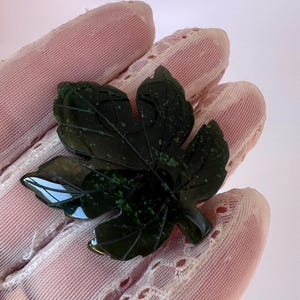 Puede incluir: Un broche en forma de hoja verde oscuro con vetas detalladas. El broche está hecho de un material pulido, posiblemente jade. La hoja se sostiene sobre un fondo rosa y con encaje.
