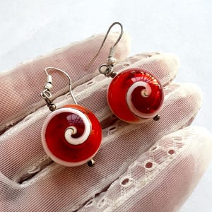 May include: A pair of red and white swirl earrings. Each earring features a round, glass bead with a red background and a white spiral design. The earrings have silver-colored ear wires.