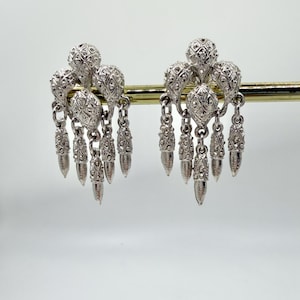 May include: A pair of silver-coloured dangle earrings. Each earring has a cluster of textured, spherical shapes connected by chains to elongated, teardrop-shaped dangles. The earrings are displayed against a white background.