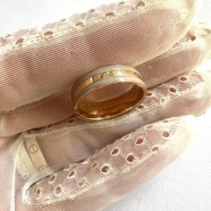 May include: A gold ring with a light gray band and the name "DANIEL" in gold lettering. The ring is held against a backdrop of a pink, lace-trimmed fabric.