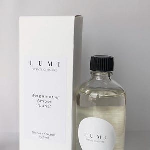 May include: A clear glass diffuser bottle with a black cap and a white label with the text "LUMI SCENTS CHESHIRE". Beside it is a white box with the same branding and the scent description "Bergamot & Amber 'Luna'". The bottle contains 100ml of liquid.