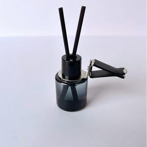 May include: A black car air freshener with a dark grey glass bottle. Two black reeds are inserted into the bottle. A black clip is attached for easy placement in a vehicle.
