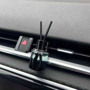 May include: A car air freshener with a black cap and two black reeds. The glass bottle has a dark, smoky appearance. The freshener is placed on a car's dashboard near the air vents.