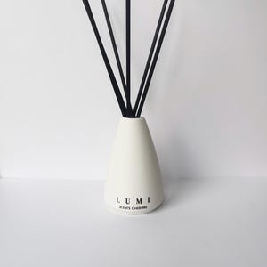 May include: A white ceramic essential oil diffuser with black reeds. The diffuser has a teardrop shape and features the text "LUMI SCENTS CHESHIRE" in black. The reeds are arranged in a fan shape, creating a minimalist aesthetic.