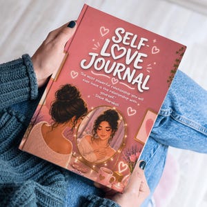 May include: A self-love journal with a pink cover featuring the words "SELF LOVE JOURNAL" in white script. The cover art includes an illustration of a woman looking in a mirror, surrounded by hearts and decorative elements. The journal is held in a person's hands.