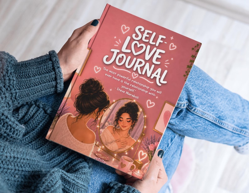 May include: A self-love journal with a pink cover featuring the words "SELF LOVE JOURNAL" in white script. The cover art includes an illustration of a woman looking in a mirror, surrounded by hearts and decorative elements. The journal is held in a person's hands.