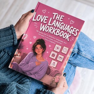 May include: A pink workbook titled "The Love Languages Workbook" with a cartoon illustration of a woman on the cover. The woman is wearing a purple outfit and sitting on a couch. The book is being held by a person wearing a blue sweater and jeans.