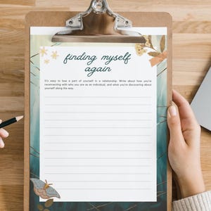 May include: A clipboard with a lined journal page featuring the text "finding myself again." The page has a decorative border with floral and butterfly illustrations. A pencil rests on the wooden surface.