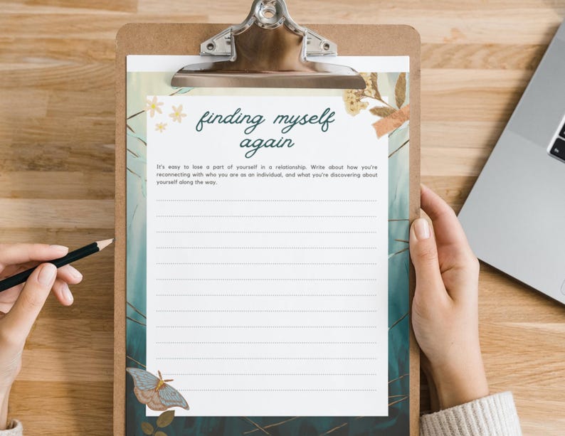 May include: A clipboard with a lined journal page featuring the text "finding myself again." The page has a decorative border with floral and butterfly illustrations. A pencil rests on the wooden surface.