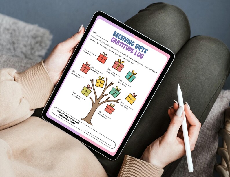May include: A digital tablet displaying a "Receiving Gifts Gratitude Log." The log features a tree with colorful gift boxes and prompts for recording gift details. A stylus rests in the hand, suggesting digital note-taking. The tablet is held on a person's lap.