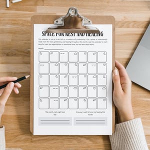 May include: A clipboard holds a printed calendar titled "SPACE FOR REST AND HEALING." The calendar features a grid of boxes with small illustrations. A hand holds a pencil, and a laptop is visible in the background.