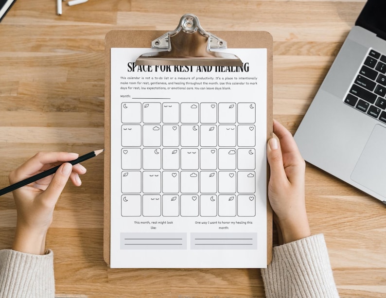 May include: A clipboard holds a printed calendar titled "SPACE FOR REST AND HEALING." The calendar features a grid of boxes with small illustrations. A hand holds a pencil, and a laptop is visible in the background.