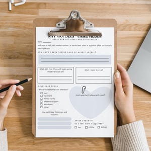 May include: A clipboard with a self-care reset worksheet. The worksheet includes sections for reflection, such as 'How have I been taking care of myself lately?' and 'Self-Care Focus'. A pencil and a laptop are also visible.