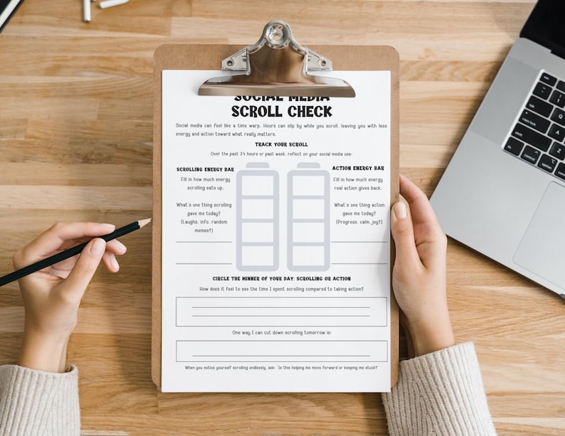 May include: A clipboard with a printed "Social Media Scroll Check" form, featuring sections for tracking energy levels and reflection. A hand holds a pencil, ready to fill out the form. A laptop is visible in the background.