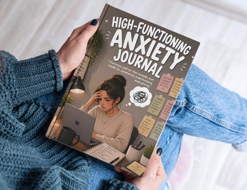 May include: A journal titled "HIGH-FUNCTIONING ANXIETY JOURNAL" is held open. The cover illustration depicts a woman at a desk with a laptop, looking stressed. The journal's text reads, "I function well on the outside, but inside I'm constantly overthinking everything."