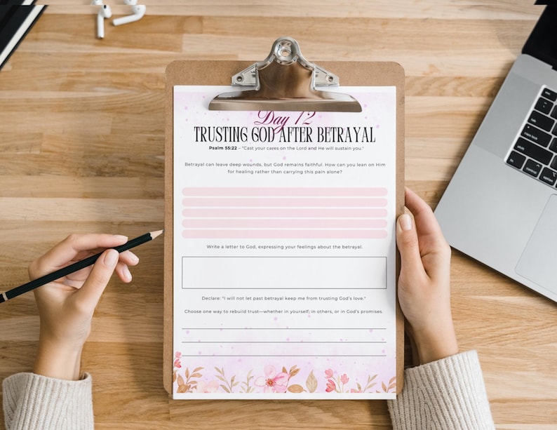 Pu&ograve; includere: Un appunti con un foglio di lavoro stampato intitolato "Trusting God After Betrayal". Il foglio di lavoro ha linee rosa e accenti floreali. Una mano tiene una matita, pronta per scrivere. Un laptop e auricolari sono sullo sfondo.