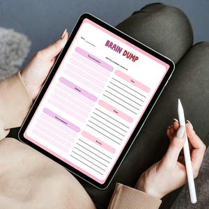 May include: A digital tablet displaying a "Brain Dump" template with pink and purple sections for identifying emotions. The tablet is held by a person, with a white stylus resting in their hand. The text on the screen is clear and readable.