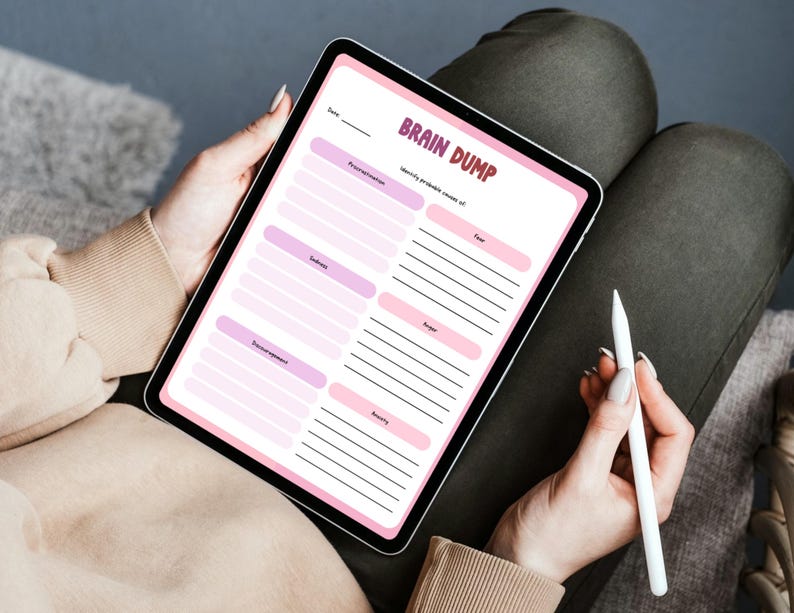 May include: A digital tablet displaying a "Brain Dump" template with pink and purple sections for identifying emotions. The tablet is held by a person, with a white stylus resting in their hand. The text on the screen is clear and readable.