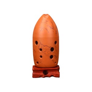 Op de afbeelding: Een oranje ocarina met donkerbruine marmering en verschillende vingergaten. Het keramische blaasinstrument wordt gepresenteerd op een kleine, roodbruine houten standaard. De ocarina heeft een afgeronde, kogelachtige vorm.