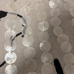 Può includere: Fili di dischi di conchiglia bianchi, rotondi e iridescenti sono collegati da piccoli anelli di metallo. I dischi sono disposti in file verticali, creando un elemento decorativo scintillante. Lo sfondo è un tessuto neutro con un motivo sottile.