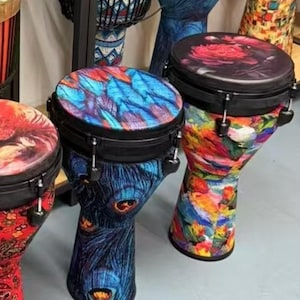 Op de afbeelding: Een verzameling kleurrijke djembé-drums met unieke ontwerpen. De drums hebben levendige patronen, waaronder pauwenveermotieven, bloemenprints en abstracte kunst. De drums worden tentoongesteld op planken en een grijs oppervlak.