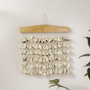 Op de afbeelding: Een wanddecoratie met rijen crèmekleurige, schelpachtige objecten die aan een natuurhouten hanger zijn bevestigd. De schelpen hebben donkere accenten en zijn geregen aan dunne, transparante koorden.