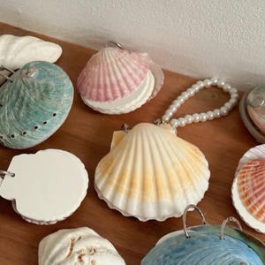 May include: A collection of seashell-shaped items in various colors, including white, pink, blue, and yellow. Some shells have a pearl beaded handle. The shells are arranged on a wooden surface.