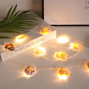 Könnte beinhalten: Eine Lichterkette mit warmweißen LEDs und Muscheldekorationen. Die Muscheln sind in verschiedenen Farben gehalten, darunter Weiß, Orange und Braun. Die Lichterkette liegt auf einer weißen Oberfläche. Der Text "Audrey Hepburn" ist sichtbar.