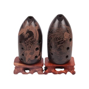 Può includere: Due ocarine marrone scuro con disegni di draghi e fenici, appoggiate su supporti di legno. Le ocarine hanno più fori per le dita e una forma arrotondata. Il drago e la fenice sono incisi in un colore marrone più chiaro.