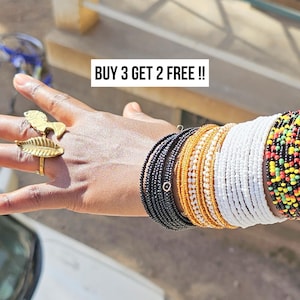 May include: A hand displays a collection of beaded bracelets in black, gold, white, and multicolored patterns. The hand also wears two gold rings, one with a leaf design and the other with a lion design. Text in the image reads: BUY 3 GET 2 FREE !!