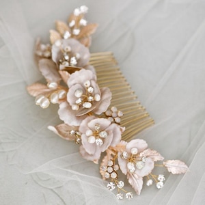 Könnte beinhalten: Ein goldfarbener Haarkamm, verziert mit zarten, rosafarbenen Stoffblüten, Kunstperlen und funkelnden Kristallen. Das Blumenarrangement umfasst detaillierte Blätter und Kristallcluster, wodurch ein elegantes Accessoire für besondere Anlässe entsteht.