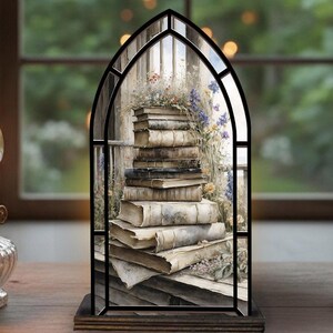 Op de afbeelding: Een decoratief glas-in-loodpaneel met een stapel oude boeken met bloemen. Het paneel is geplaatst in een zwart boogvormig frame en staat op een houten voet. De achtergrond toont een raam met een vage buitenscène.