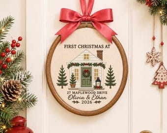 May include: A round wooden embroidery hoop containing fabric with a stitched house illustration, green trees, and the text FIRST CHRISTMAS AT 27 MAPLEWOOD DRIVE Olivia & Ethan 2026. A red satin ribbon bow is tied at the top for hanging.