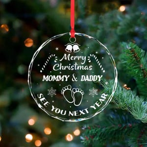 Op de afbeelding: Een heldere, ronde kerstversiering met de tekst "Merry Christmas Mommy & Daddy See You Next Year". De versiering heeft een rood lint om op te hangen en toont babyvoetjes en sneeuwvlokken.