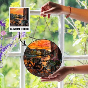 Puede incluir: Una obra de arte redonda de vidrieras que representa una puesta de sol sobre un canal con bicicletas. La obra de arte incluye el texto "Best Trip Ever 2025" y "Custom Photo". Se cuelga de una cadena.