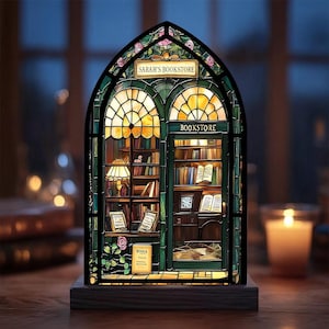 May include: A stained glass bookstore lamp with the text "SARAH'S BOOKSTORE" and "BOOKSTORE". The lamp has a wooden base, a green and gold frame, and a detailed interior with books and a lamp. The design includes pink roses.