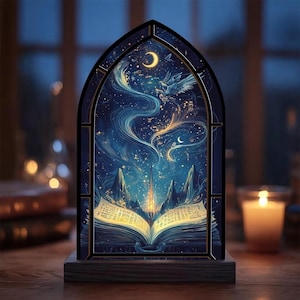May include: A decorative stained glass panel depicting a celestial scene with a crescent moon, stars, and a flowing, ethereal design. The panel is set in a dark frame and rests on a wooden base. An open book with illuminated pages is at the bottom.