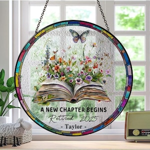 May include: Circular stained glass window art featuring a floral design and a butterfly. The text reads "A New Chapter Begins Retired 2025 - Taylor -". The window has a mosaic border and is suspended by a chain. The window is approximately 25 cm in diameter.