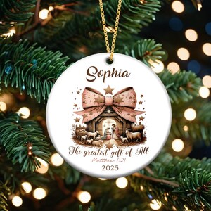 May include: A white ceramic Christmas ornament with a gold chain. The ornament features a nativity scene with a pink bow, the name "Sophia", and the text "The greatest gift of All" and "2025".