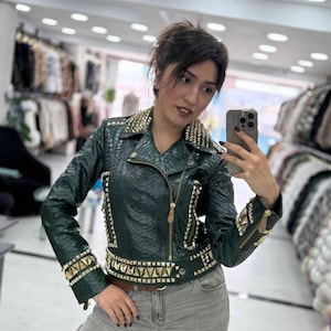 May include: Dark green leather jacket with gold studs and zips. The jacket has a snakeskin texture and a brown belt. The collar and cuffs are heavily studded. The model is wearing grey jeans.
