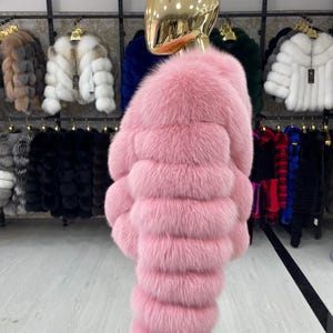 May include: A vibrant pink fur coat displayed on a mannequin. The coat features horizontal bands of plush fur, creating a textured appearance. Other fur coats in various colors are visible in the background, suggesting a collection of outerwear.