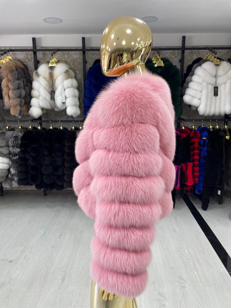 May include: A vibrant pink fur coat displayed on a mannequin. The coat features horizontal bands of plush fur, creating a textured appearance. Other fur coats in various colors are visible in the background, suggesting a collection of outerwear.