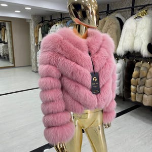 May include: A pink fur jacket with a round neckline and long sleeves is displayed on a mannequin. The jacket has a chevron design and a black tag. The mannequin is gold-colored and is in a clothing store.
