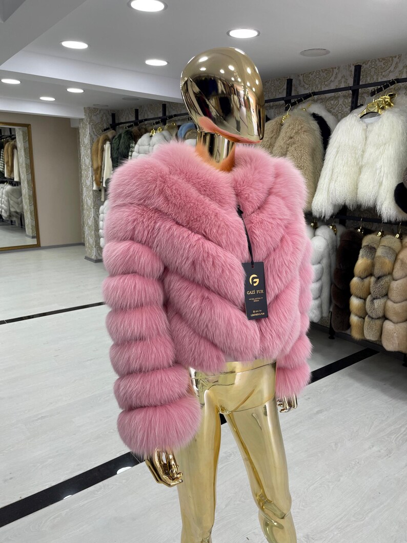 May include: A pink fur jacket with a round neckline and long sleeves is displayed on a mannequin. The jacket has a chevron design and a black tag. The mannequin is gold-colored and is in a clothing store.