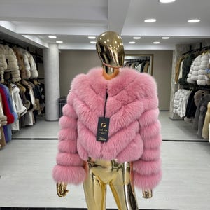 May include: A pink fur jacket with a chevron design is displayed on a mannequin with a gold head and legs. The jacket has a zip closure and long sleeves. A tag with the brand name is attached to the jacket.