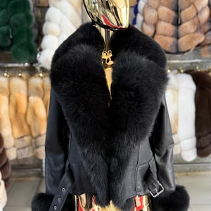 Puede incluir: Chaqueta de cuero negro con un gran cuello y puños de piel negra y esponjosa. La chaqueta tiene un cinturón en la cintura y se exhibe en un maniquí dorado. Se ven otros abrigos de piel en el fondo.