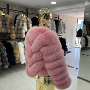 May include: A pink fur jacket with a cropped design is displayed on a mannequin. The jacket has a textured, horizontal pattern and a plush appearance. Other fur jackets in various colors are visible in the background.