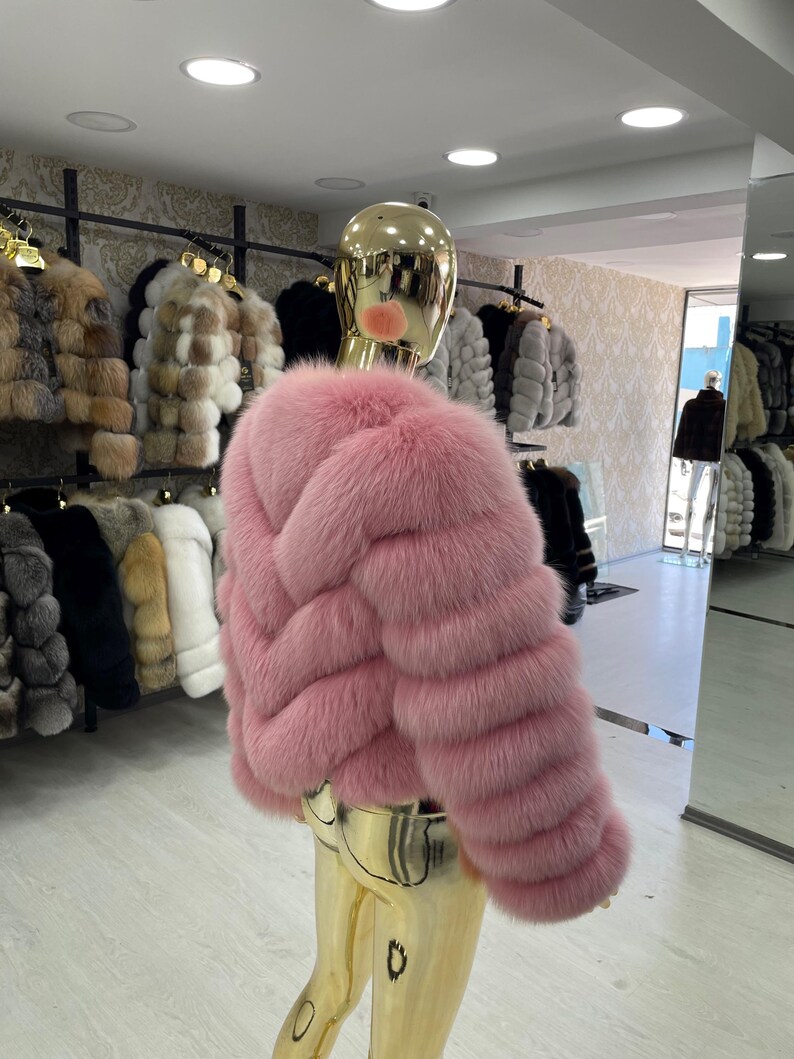 May include: A pink fur jacket with a cropped design is displayed on a mannequin. The jacket has a textured, horizontal pattern and a plush appearance. Other fur jackets in various colors are visible in the background.
