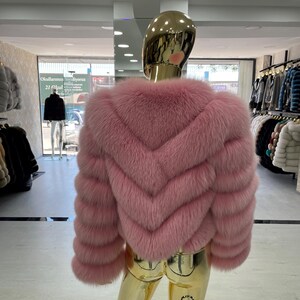 May include: A pink fur jacket with a chevron design on the back. The jacket is displayed on a gold mannequin in a clothing store. The sleeves are long and the fur appears soft and plush. The jacket is a light shade of pink.